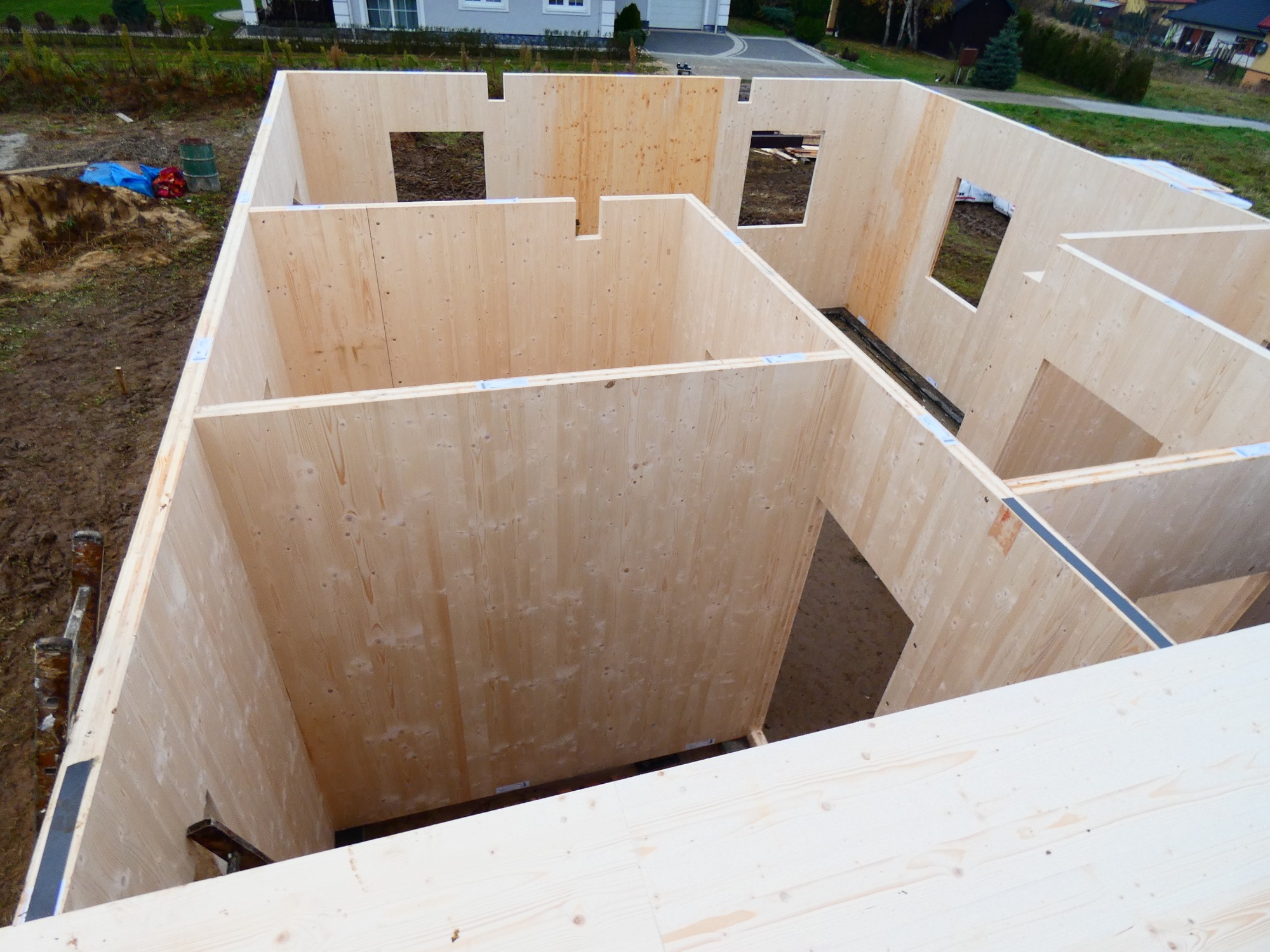 Wnętrze surowego stanu domu z drewnianą konstrukcją szkieletową, widok z góry na ściany działowe i otwory okienne, z widocznymi elementami montażowymi i otoczeniem budowy.