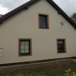 Ściana boczna domu po remoncie z beżową elewacją i brązowymi obramowaniami okien, widoczne skrzynki elektryczne i częściowo rusztowanie.