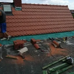 Montaż czerwonej dachówki karpiówki na dachu z kominem; widoczne narzędzia, zielona membrana i drewniane listwy.