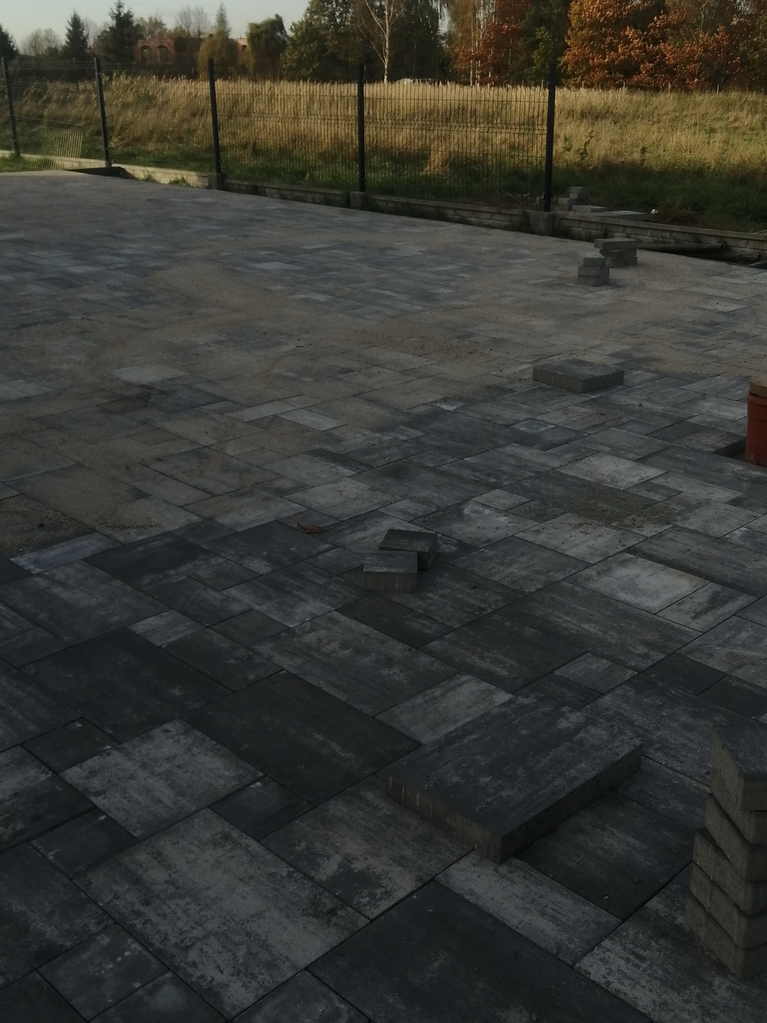 Układanie szarej kostki brukowej na posesji, widoczne stosy kostki i fragmenty powierzchni jeszcze bez wyłożenia. W tle ogrodzenie z siatki i zieleń.