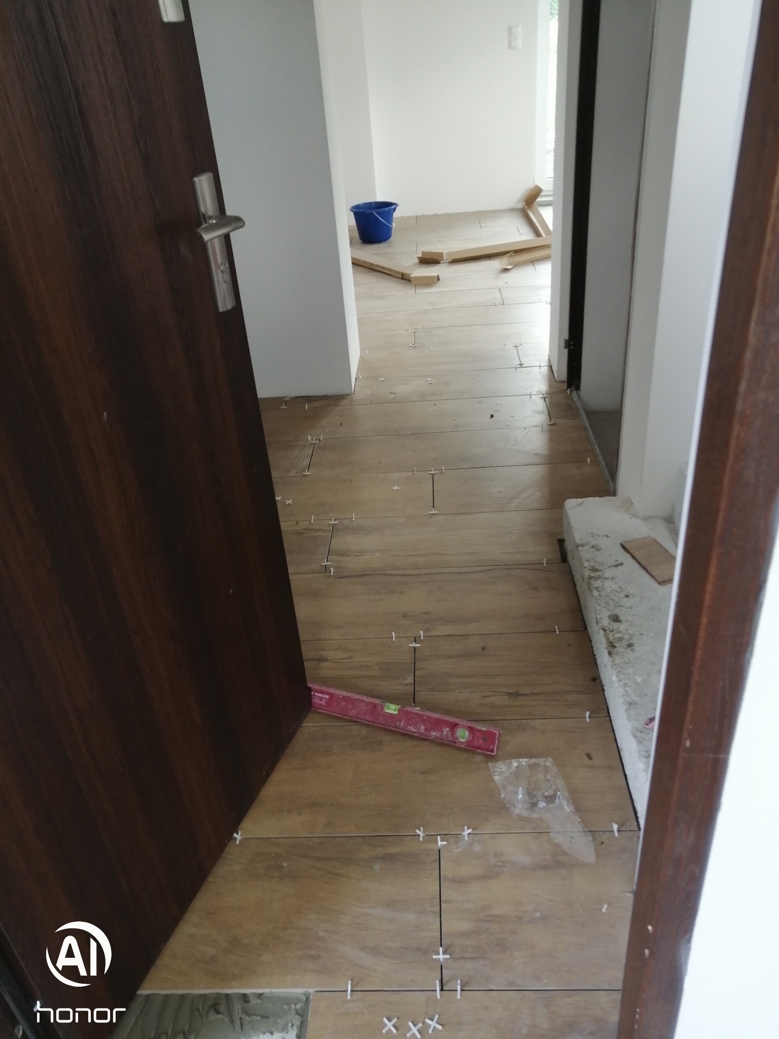 Układanie płytek podłogowych imitujących drewno w korytarzu, widoczna poziomica i krzyżyki dystansowe. Częściowo widoczna ościeżnica i drzwi.