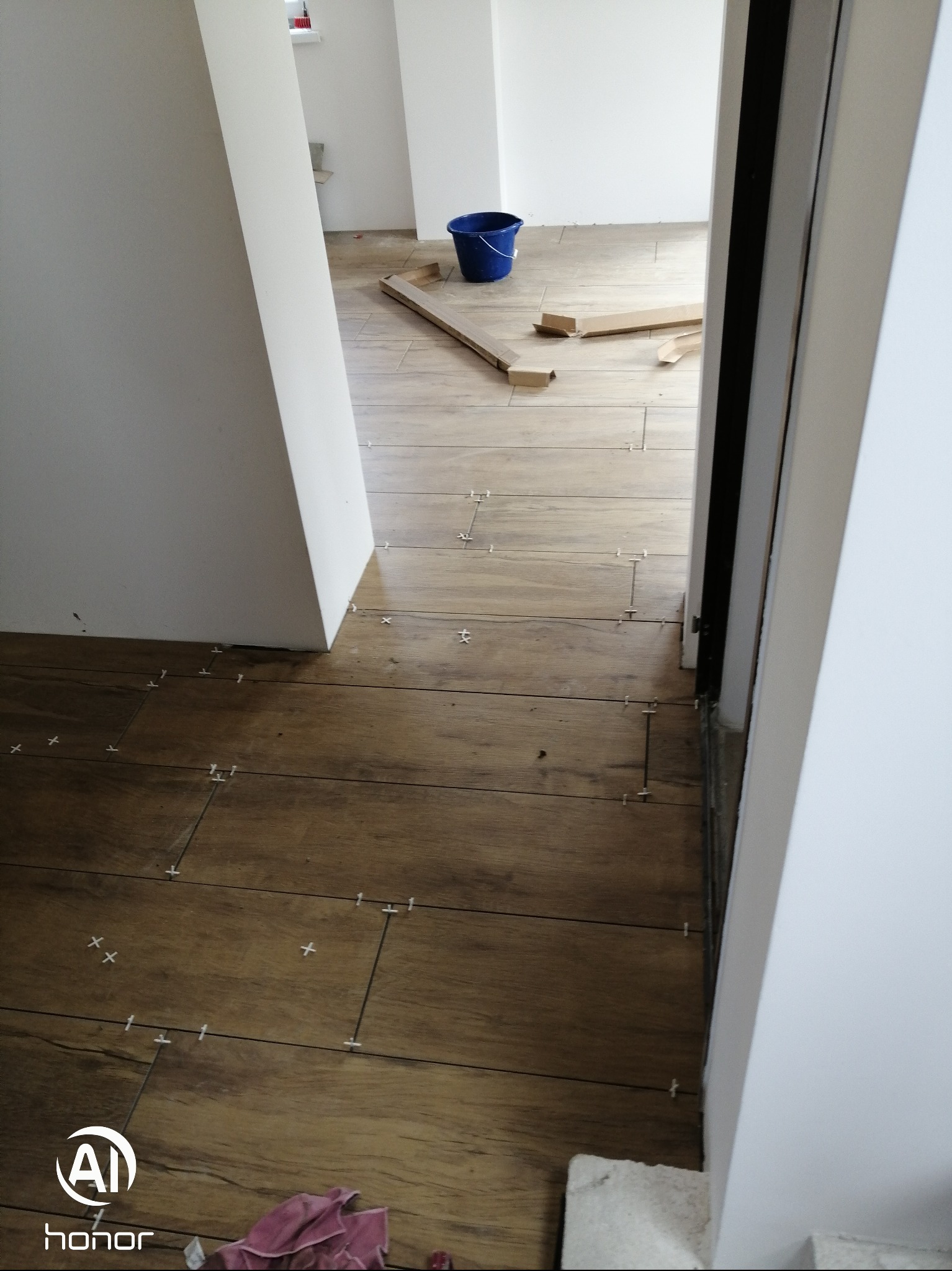 Układanie podłogi z płytek imitujących drewno w jasnym wnętrzu. Widoczne krzyżyki dystansowe i niebieskie wiadro w tle. Praca w toku, z perspektywy wejścia do pomieszczenia.