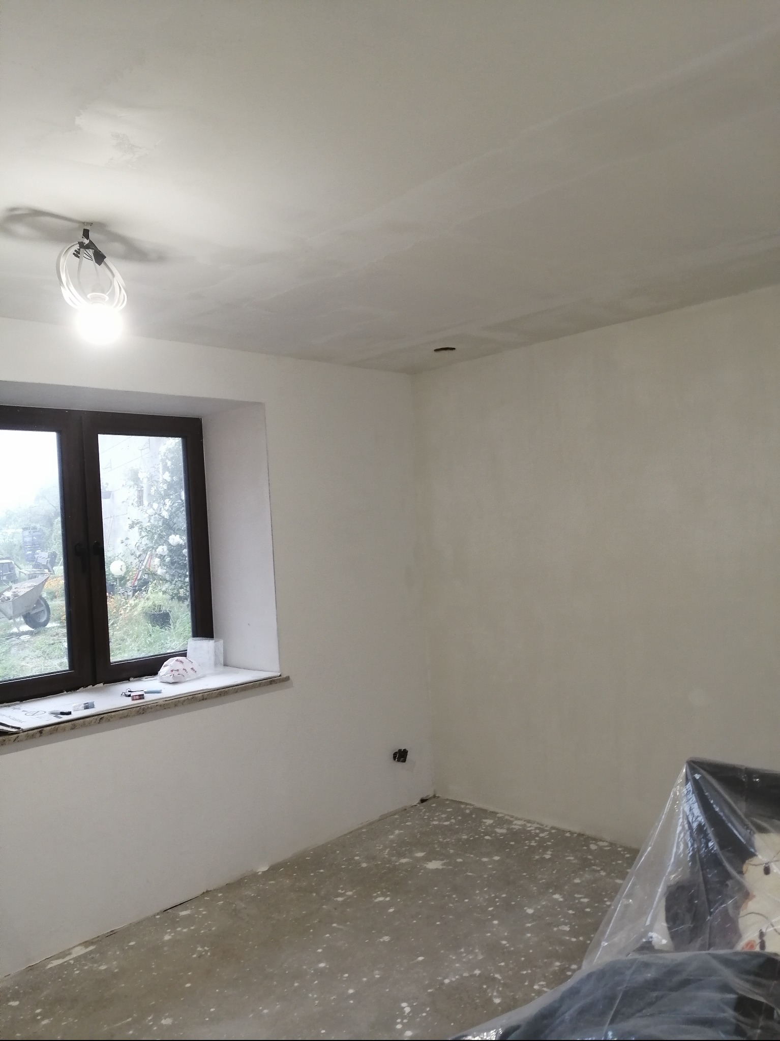 Wnętrze pokoju po szpachlowaniu ścian i sufitu, widoczne gładkie powierzchnie przygotowane do malowania. Okno z widokiem na zewnątrz, pod oknem akcesoria malarskie.