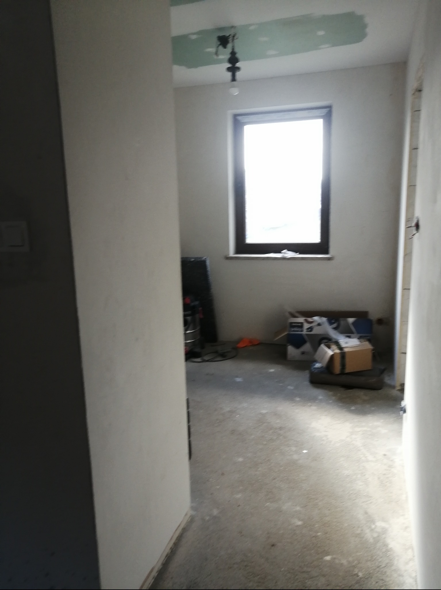 Wnętrze w trakcie remontu: surowy pokój z oknem, widoczne ślady szpachlowania na suficie, kartony na podłodze. Ujęcie z perspektywy wejścia, z widoczną częścią korytarza.
