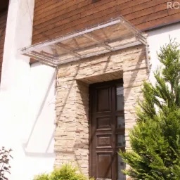 Zadaszenie nad wejściem do domu z kamienną elewacją, konstrukcja ze stali nierdzewnej i przezroczystym wypełnieniem, widoczna roślinność.