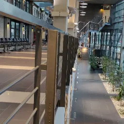 Wnętrze budynku z metalową balustradą o pionowych elementach z napisem, widok na schody, rośliny i siedzenia w hali.