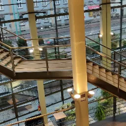 Wielopoziomowe schody metalowe z drewnianymi stopniami i balustradami ze stali nierdzewnej, widoczne z góry, z dużymi oknami w tle ukazującymi miejski krajobraz.