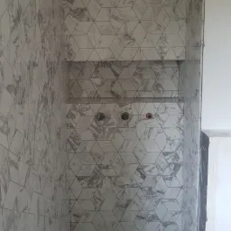 Ściany i podłoga w łazience wyłożone płytkami w geometryczny wzór imitujący marmur, z wnękami w ścianie przygotowanymi pod półki i widocznymi instalacjami hydraulicznymi.