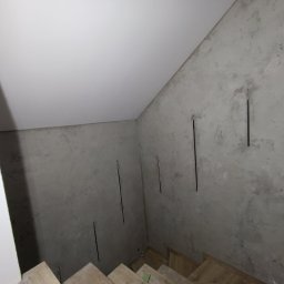 Twoje-Remonty Tomasz Kucicki - Ściana z szarą gładzią dekoracyjną w trakcie wykańczania, widoczne pionowe bruzdy po szpachlowaniu, fragment schodów z drewnianymi stopniami, biały sufit.
