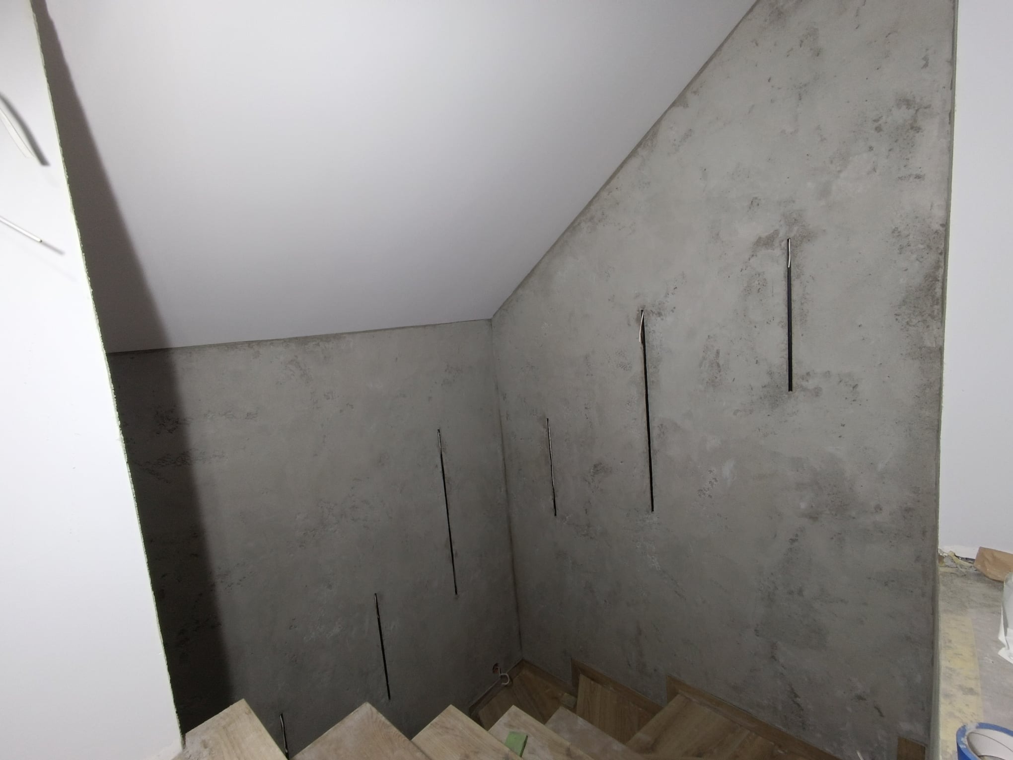 Ściana z szarą gładzią dekoracyjną w trakcie wykańczania, widoczne pionowe bruzdy po szpachlowaniu, fragment schodów z drewnianymi stopniami, biały sufit.