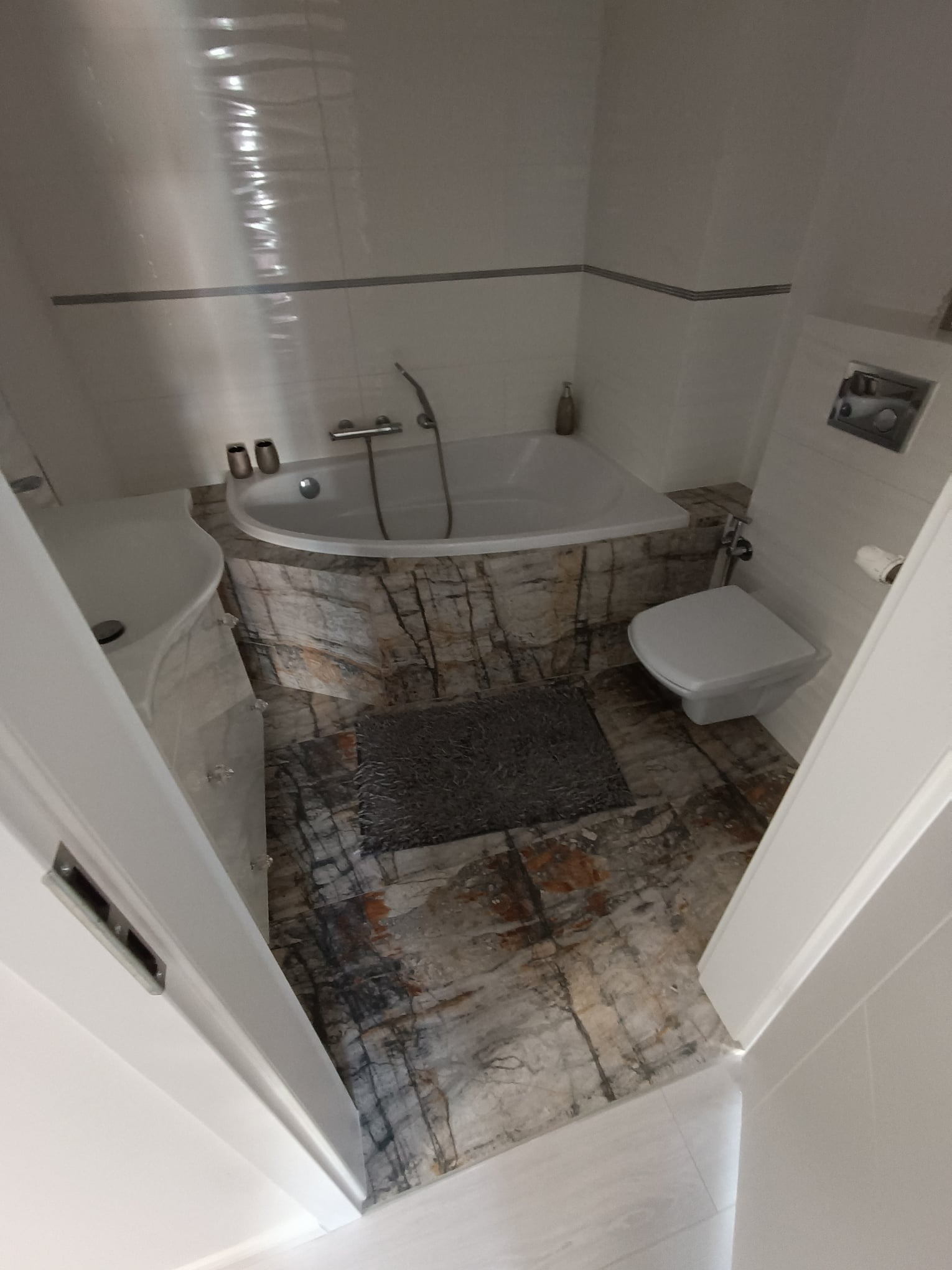 Nowoczesna łazienka z wanną w zabudowie z motywem marmuru, umywalką na postumencie, podwieszaną toaletą i podłogą z płytek imitujących kamień.