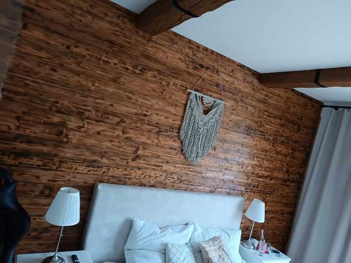 Sypialnia z drewnianą ścianą z desek, dekoracją makrama i beżowym zagłówkiem łóżka, widoczne lampki nocne i fragment zasłony.