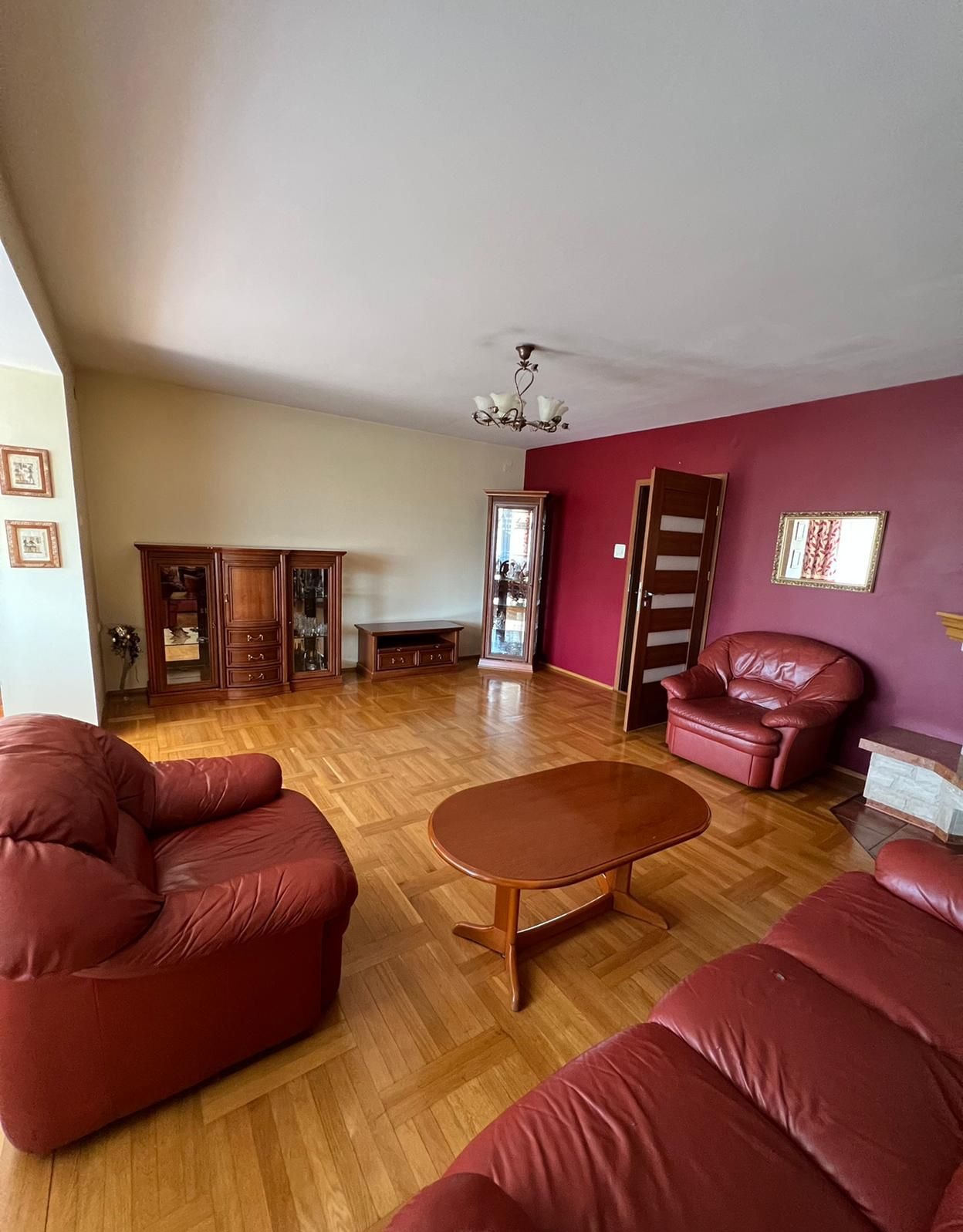 Salon po remoncie z czerwonymi ścianami, drewnianą podłogą parkietową i meblami w stylu retro, widok na otwarte drzwi.