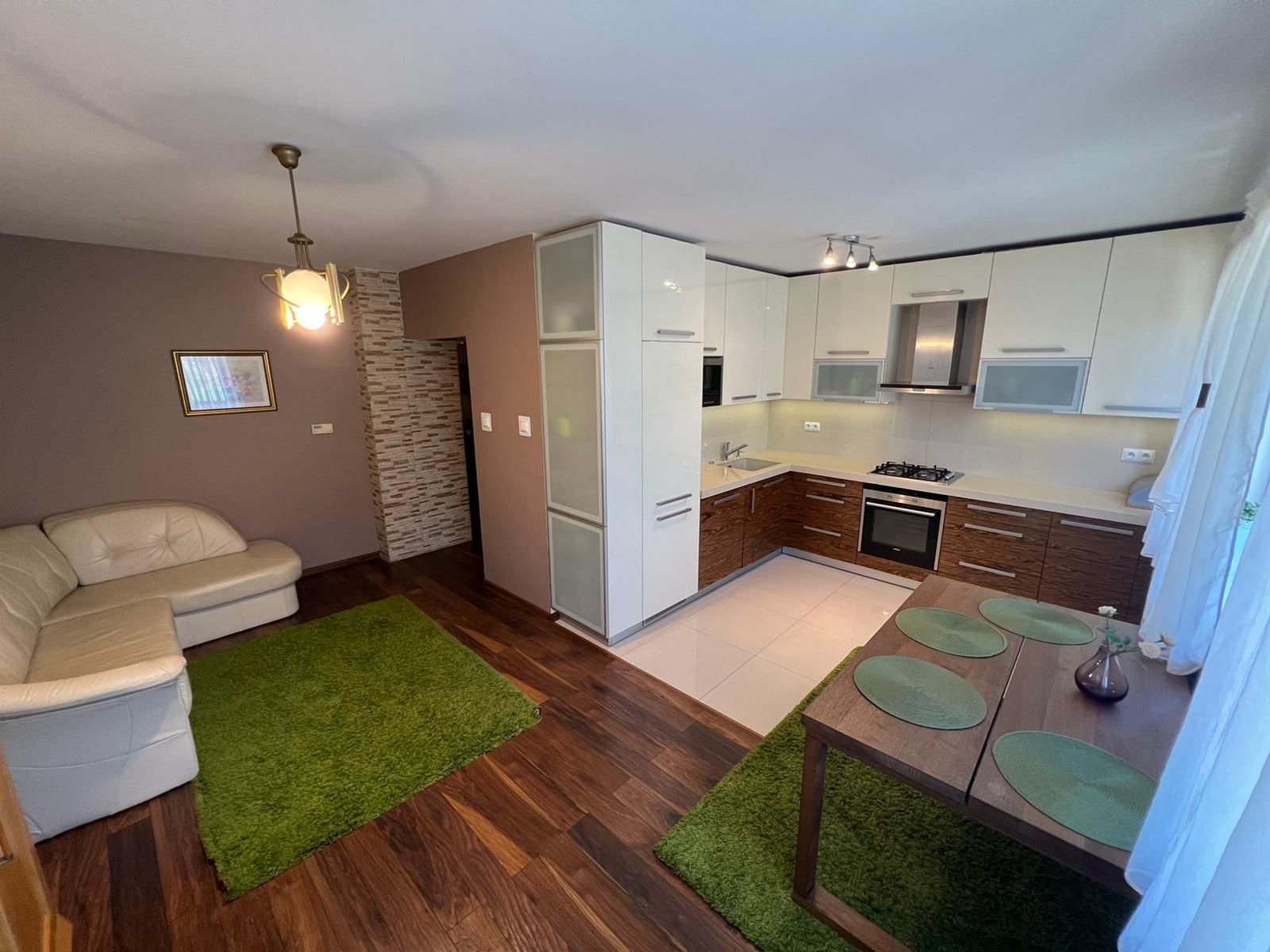 Salon z aneksem kuchennym w odcieniach brązu i bieli, z zielonymi akcentami w postaci dywanu i podkładek na stole. Widoczne elementy dekoracyjne: lampa wisząca, obraz na ścianie i kwiaty w wazonie.