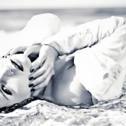 Czarno-białe zdjęcie kobiety leżącej w płytkiej wodzie, w białej koszuli, z rękami przy twarzy i ciemnym makijażem, w tle widać morze i logo fotografa.