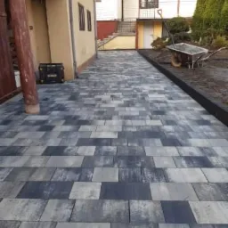 Ułożona ścieżka z szaro-niebieskiej kostki brukowej prowadząca wzdłuż domu, z widocznym wózkiem i narzędziami ogrodniczymi w tle.