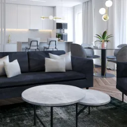 Nowoczesny salon z ciemnoniebieską sofą, marmurkowymi stolikami kawowymi, okrągłym stołem jadalnym z szarymi krzesłami oraz otwartą kuchnią w tle, całość utrzymana w jasnej kolorystyce.