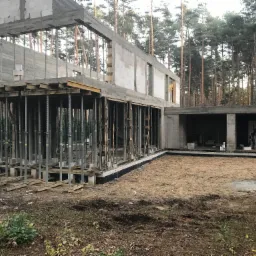 Surowy stan budynku z betonu komórkowego w otoczeniu lasu, widoczne podpory szalunkowe i fundament izolowany, teren budowy.