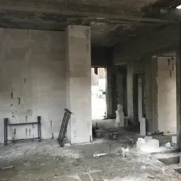 Wnętrze surowego domu murowanego w trakcie budowy, widoczne ściany z betonu komórkowego, betonowy strop, narzędzia budowlane i metalowe podpory na podłodze, przez otwór drzwiowy widoczny teren...