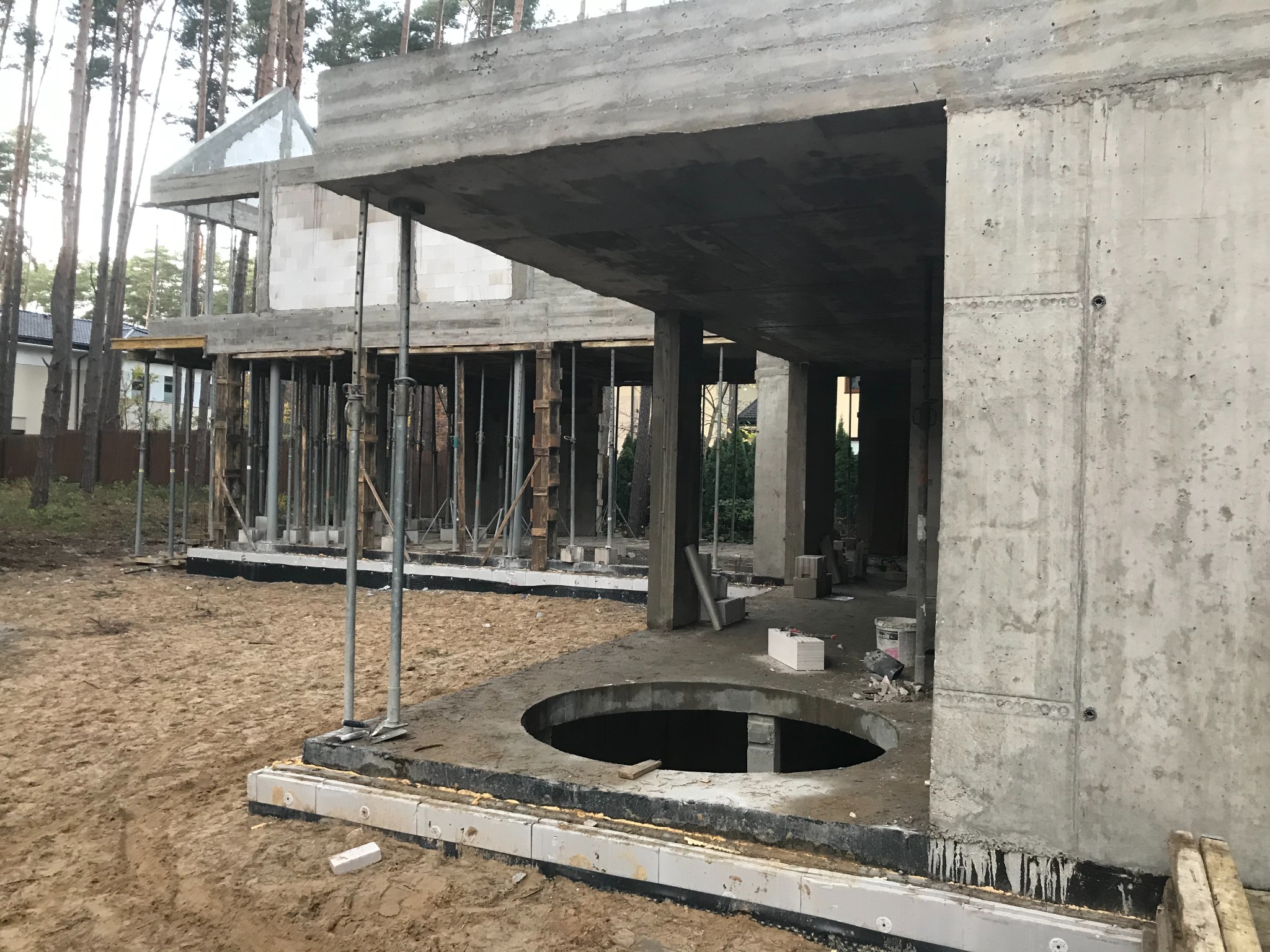 Surowy stan budynku w trakcie budowy, widoczne betonowe ściany, kolumny i stropy, zbrojenie, drewniane podpory oraz wycięty okrąg w płycie fundamentowej.