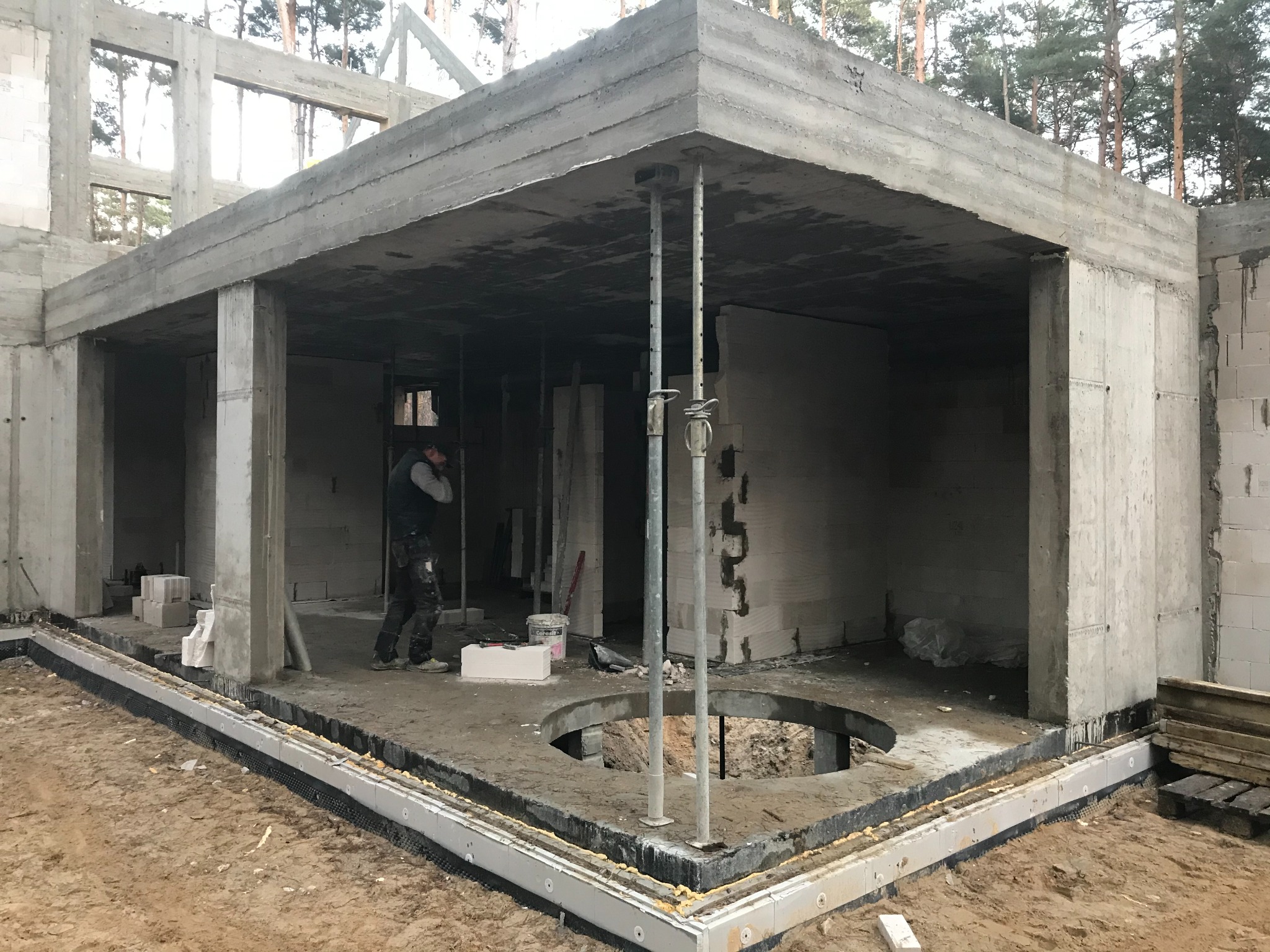 Surowy stan budynku żelbetowego z widocznymi podporami stropu, otworem w podłodze, izolacją fundamentów i pracownikiem w tle.