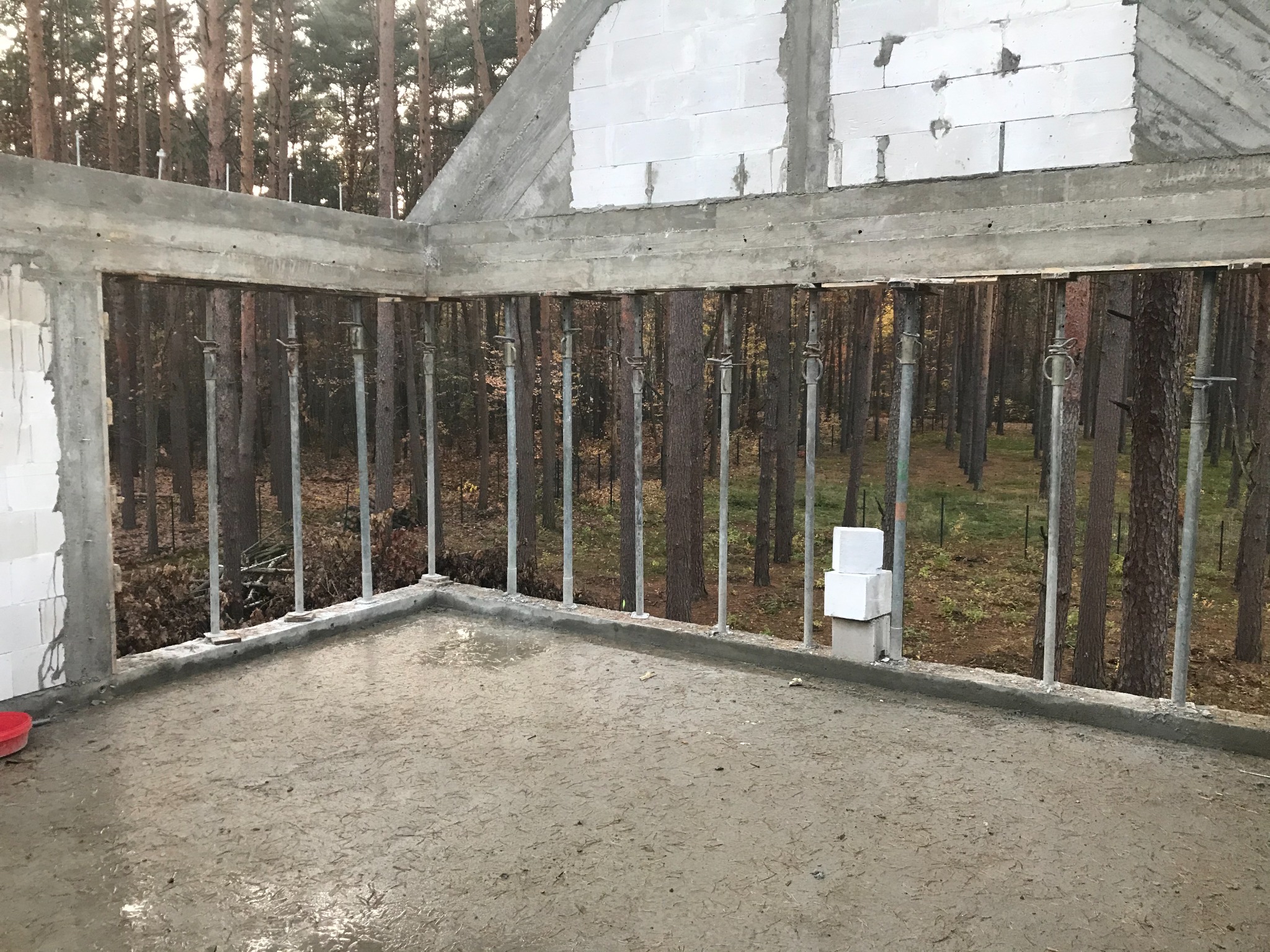 Wylany fundament z betonu zbrojonego, wsparty metalowymi stemplami na tle lasu, widoczne białe bloczki i niedokończone ściany z betonu komórkowego.