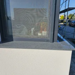 Szara elewacja z oknem i parapetem z kamienia w Kamienicy. Na parapecie kwiaty. Widoczne rusztowanie i zaparkowany samochód. Detal wykończenia.