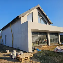 Nowoczesny dom jednorodzinny w trakcie tynkowania elewacji zewnętrznej, widoczne materiały budowlane i dwóch pracowników przy pracy.