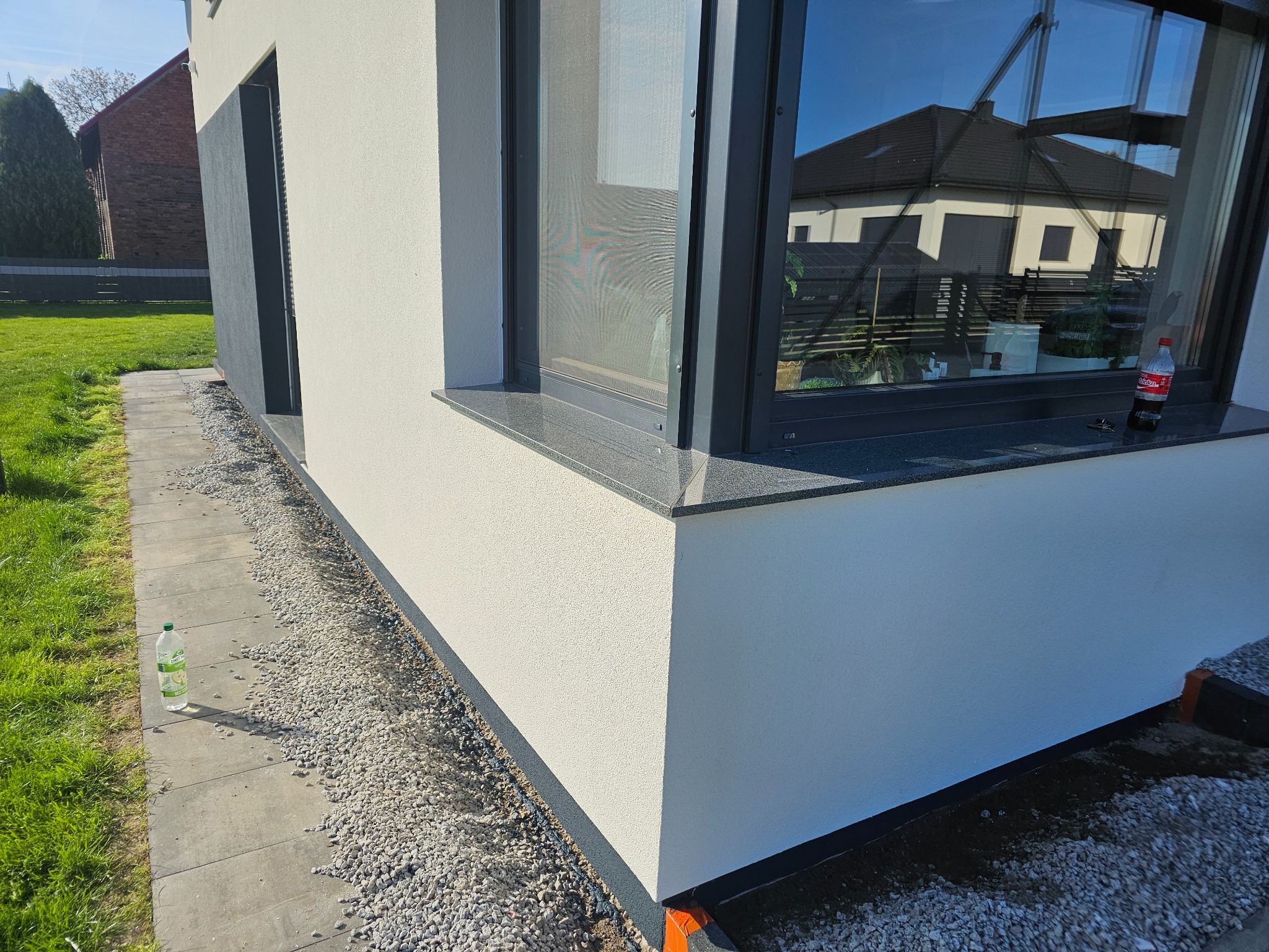 Wykończona elewacja domu z oknem. Widoczne detale: szara podmurówka, jasny tynk strukturalny, kamienny parapet, okno z moskitierą. Częściowo zasypane kamieniami.