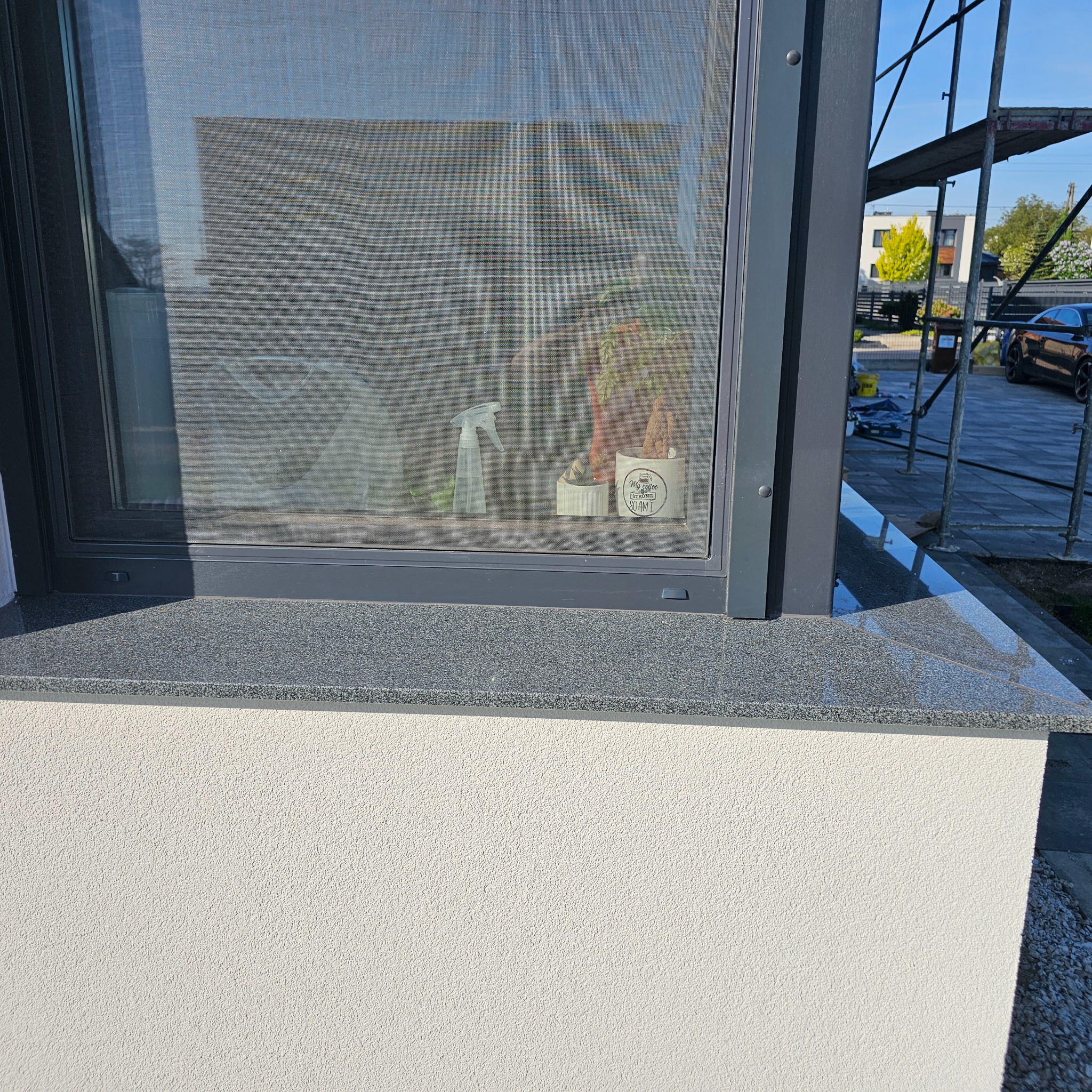 Szara elewacja z oknem i parapetem z kamienia w Kamienicy. Na parapecie kwiaty. Widoczne rusztowanie i zaparkowany samochód. Detal wykończenia.