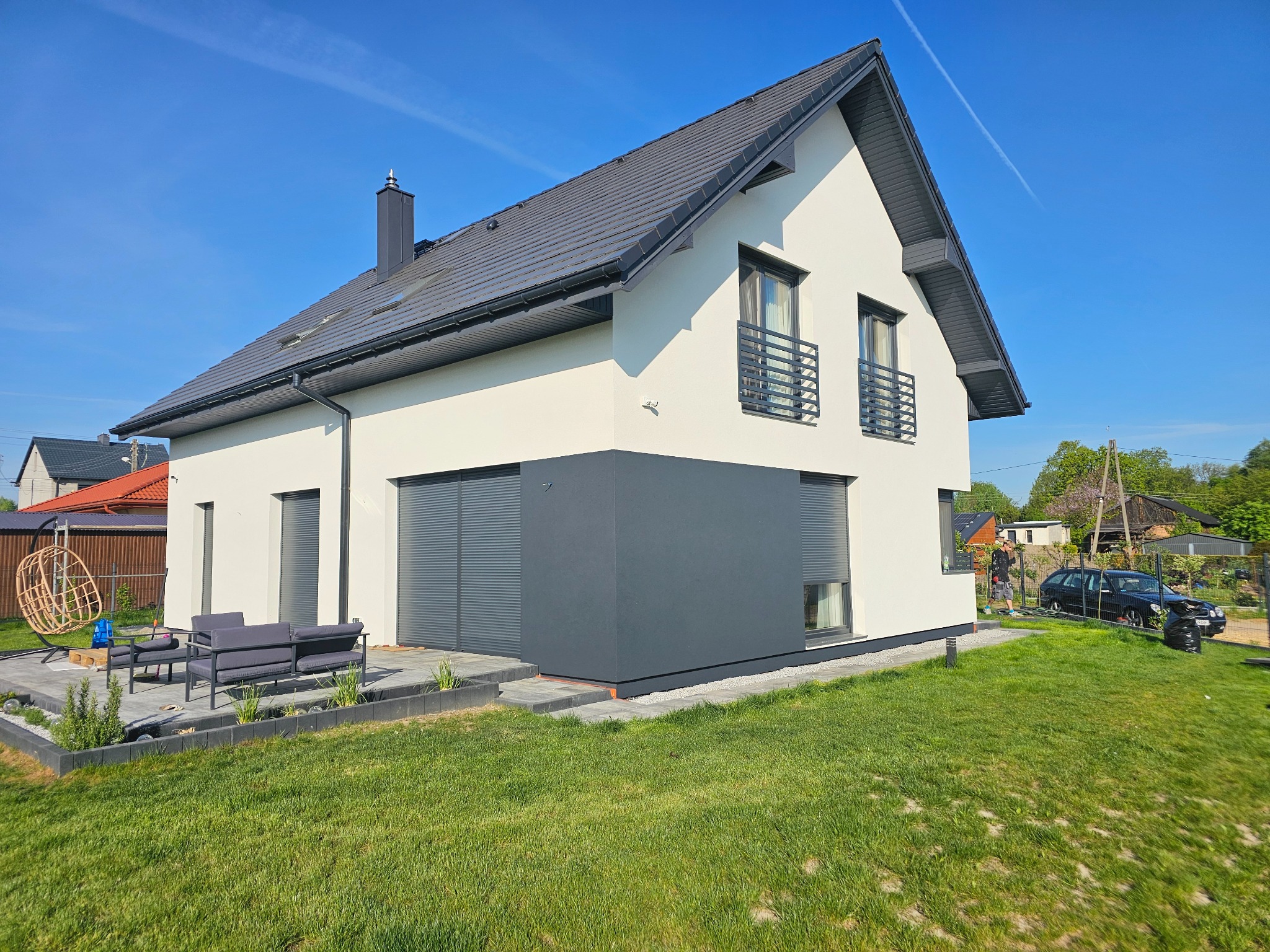 Nowoczesny dom z dwukolorową elewacją (biało-szarą), grafitowym dachem i oknami z balustradami. Widoczny taras z meblami ogrodowymi i zadbany trawnik.