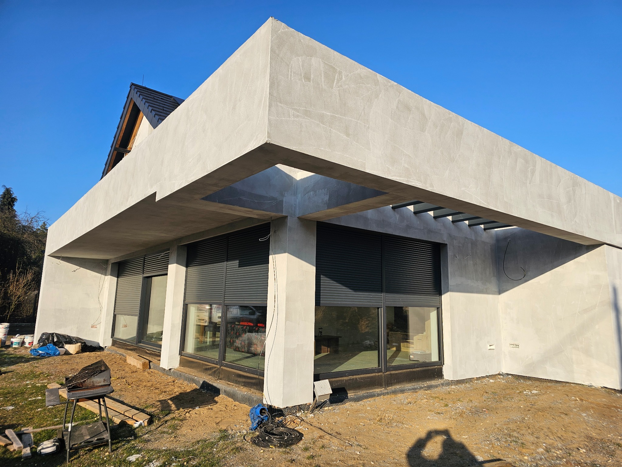 Surowy, nowoczesny dom z szarą elewacją i dużymi oknami z roletami. Widoczny fragment dachu i otoczenie w trakcie prac wykończeniowych. Minimalistyczny styl.