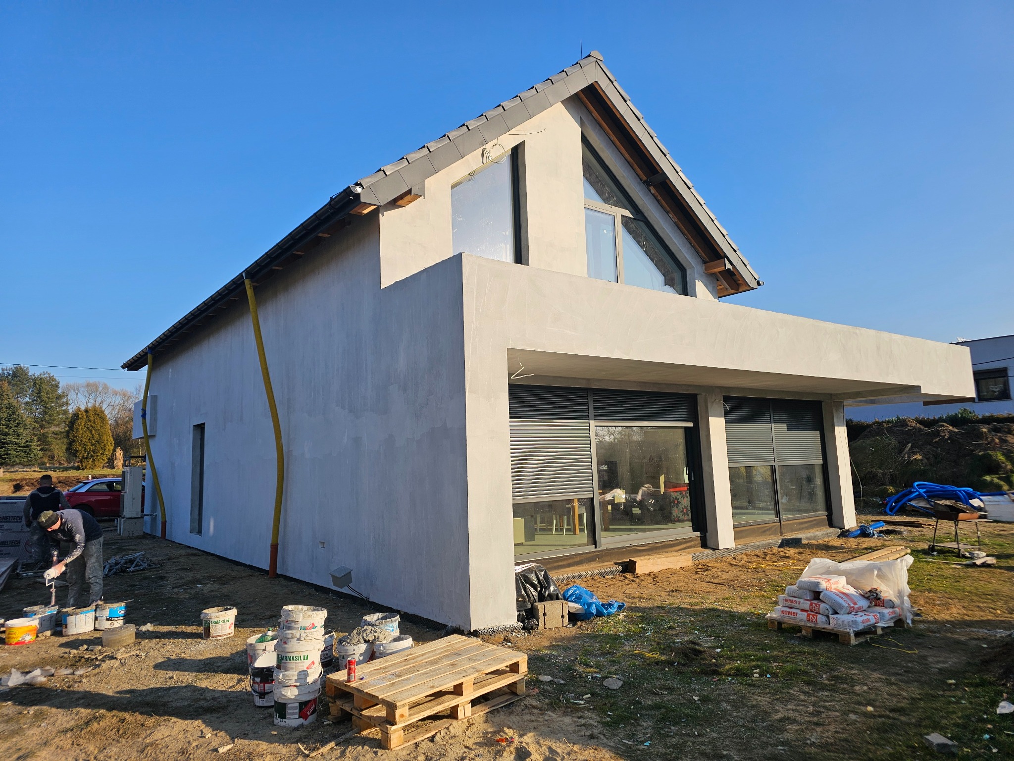 Nowoczesny dom jednorodzinny w trakcie tynkowania elewacji zewnętrznej, widoczne materiały budowlane i dwóch pracowników przy pracy.