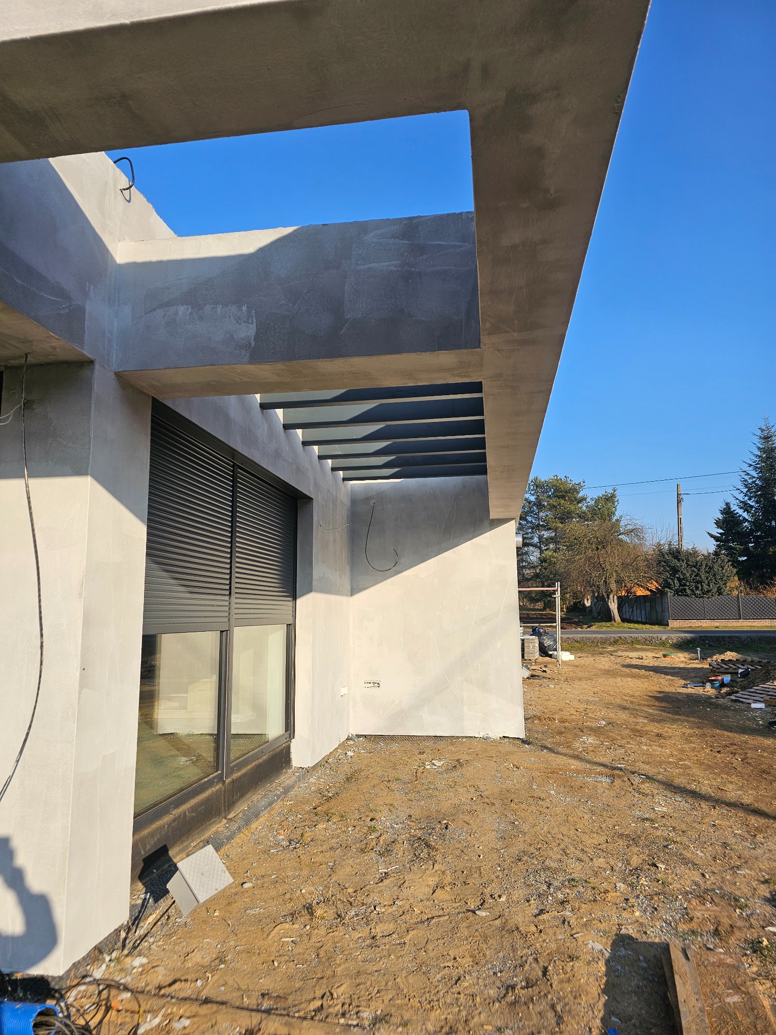 Surowa elewacja domu z szarymi roletami i betonowym zadaszeniem w trakcie prac wykończeniowych, widok z dołu na tle błękitnego nieba i fragmentu ogrodu.