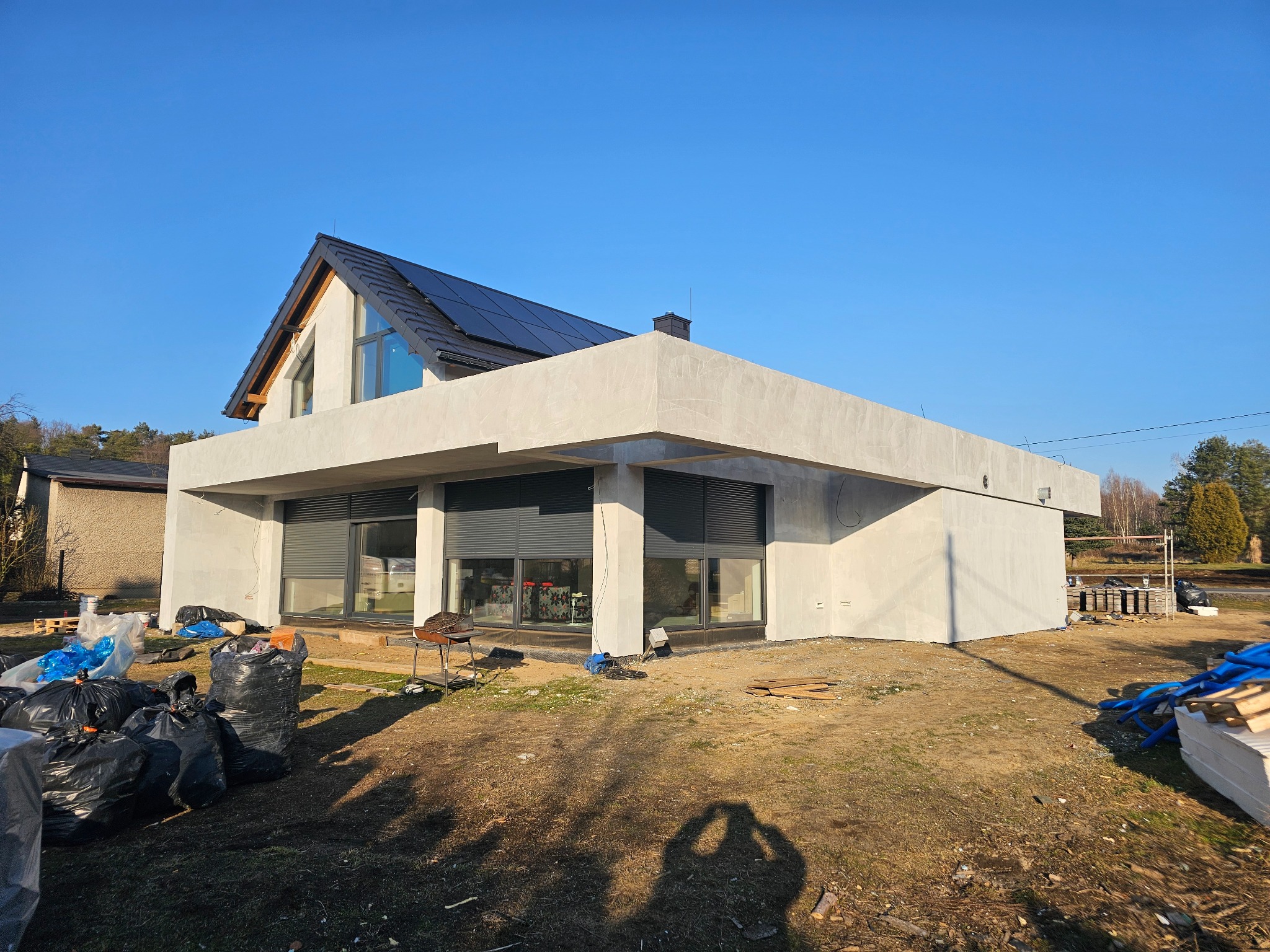 Nowoczesny dom z elewacją w trakcie tynkowania, widoczne worki z materiałami budowlanymi i narzędzia. Dach pokryty panelami słonecznymi. Błękitne niebo w tle.