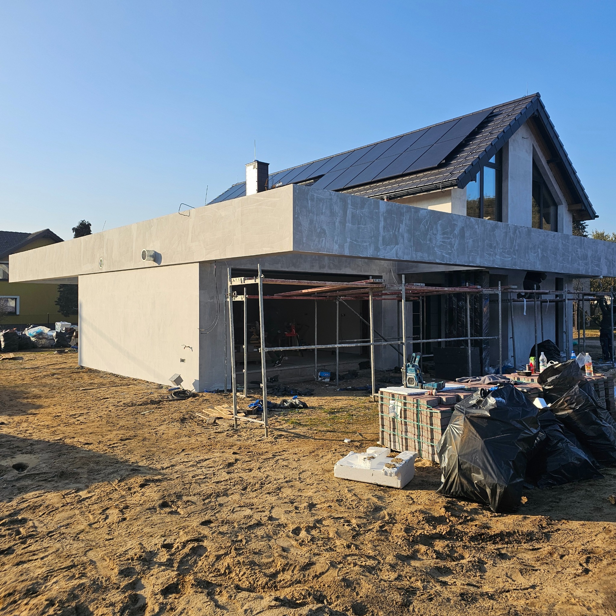 Dom jednorodzinny w trakcie tynkowania elewacji, widoczne rusztowania i materiały budowlane. Dach pokryty panelami słonecznymi. Jasne, słoneczne ujęcie.