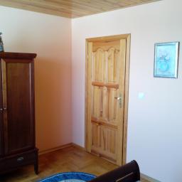 USŁUGI REMONTOWO-BUDOWLANE Radosław Kaczmarek - Jasne wnętrze pokoju z drewnianą podłogą i drzwiami, szafą z dekoracyjnym wazonem na górze oraz obrazkiem z kwiatami na ścianie, oświetlone lampą ścienną.