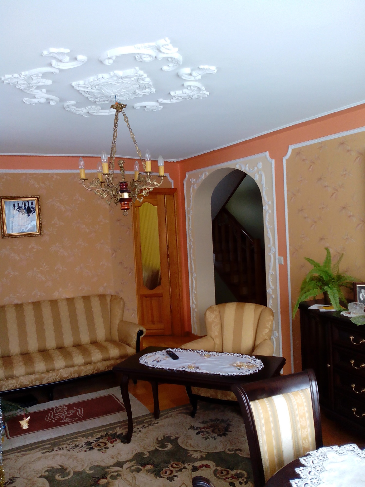 Salon w stylu klasycznym z dekoracyjnymi sztukateriami na suficie i ścianach, złocony żyrandol, meble w odcieniach beżu i brązu.