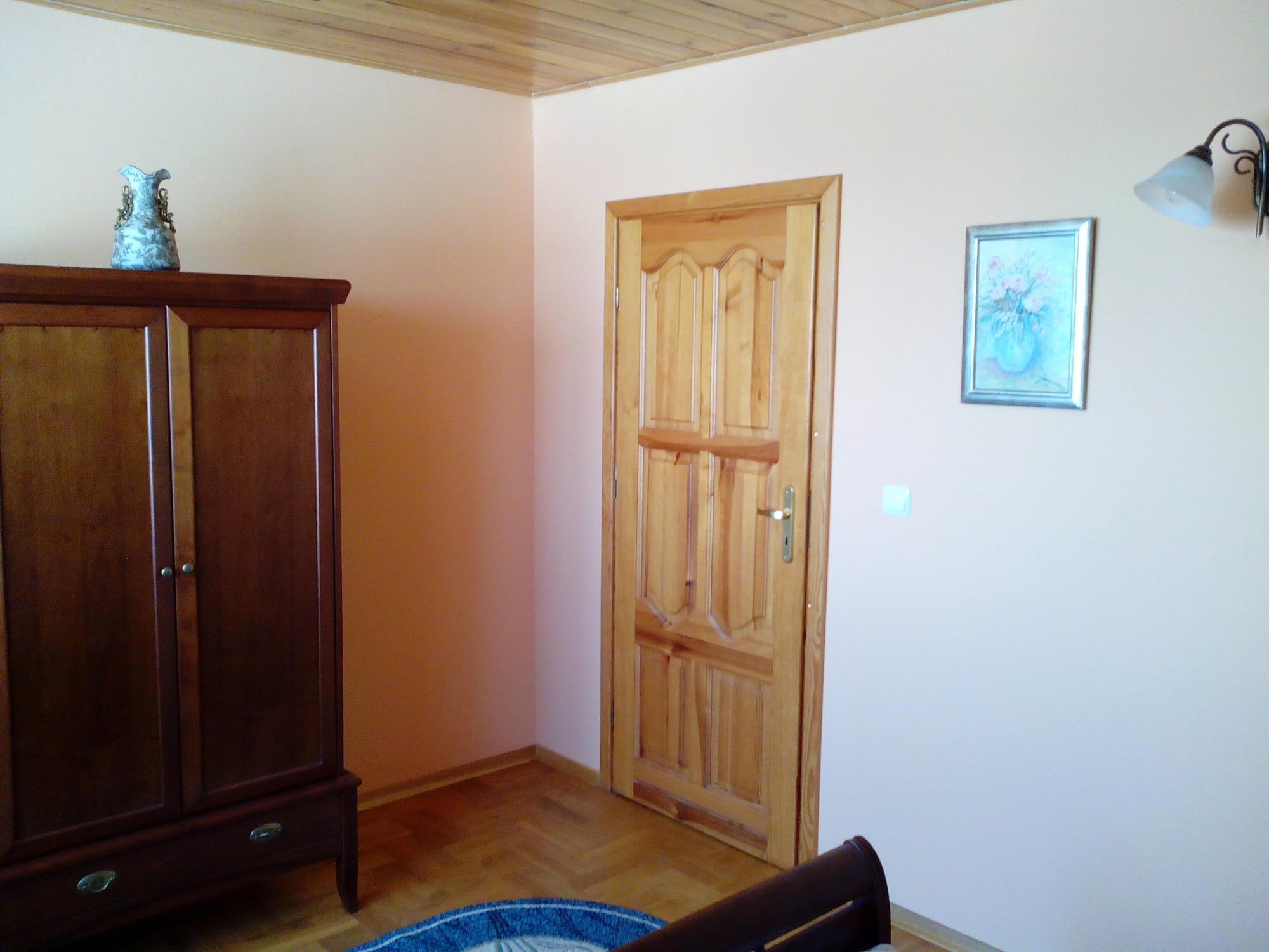 Jasne wnętrze pokoju z drewnianą podłogą i drzwiami, szafą z dekoracyjnym wazonem na górze oraz obrazkiem z kwiatami na ścianie, oświetlone lampą ścienną.