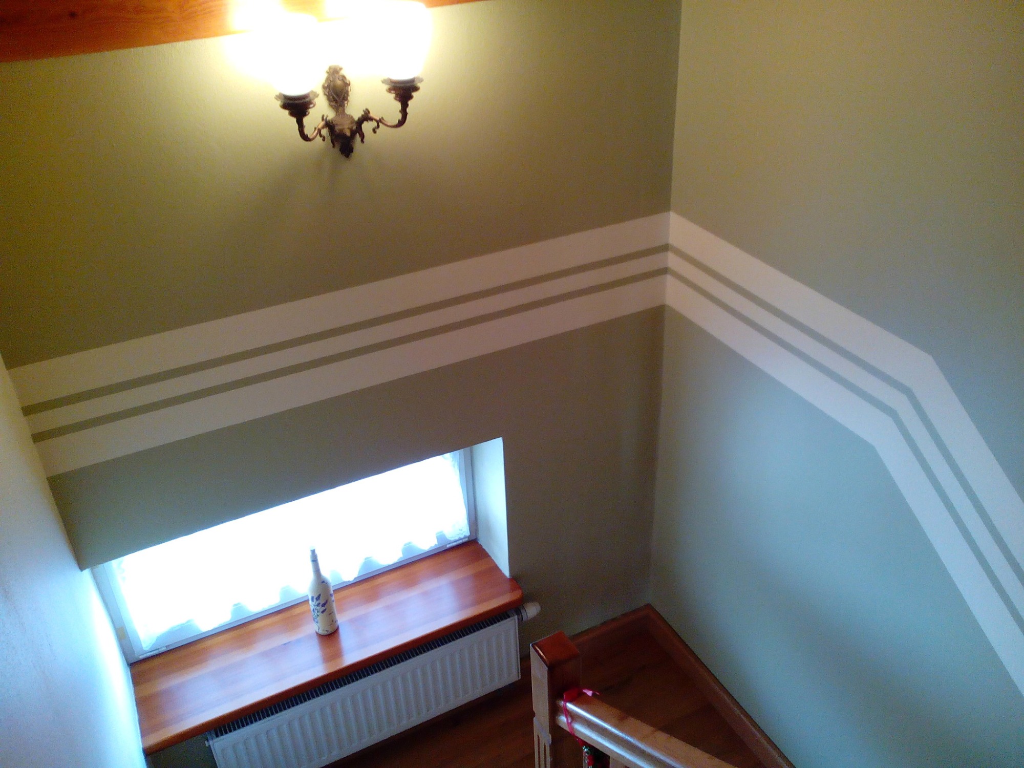 Ściana klatki schodowej pomalowana w odcieniach szarości z dekoracyjnymi, poziomymi pasami w jaśniejszym kolorze, widok z góry, na parapecie butelka z dekoracją.