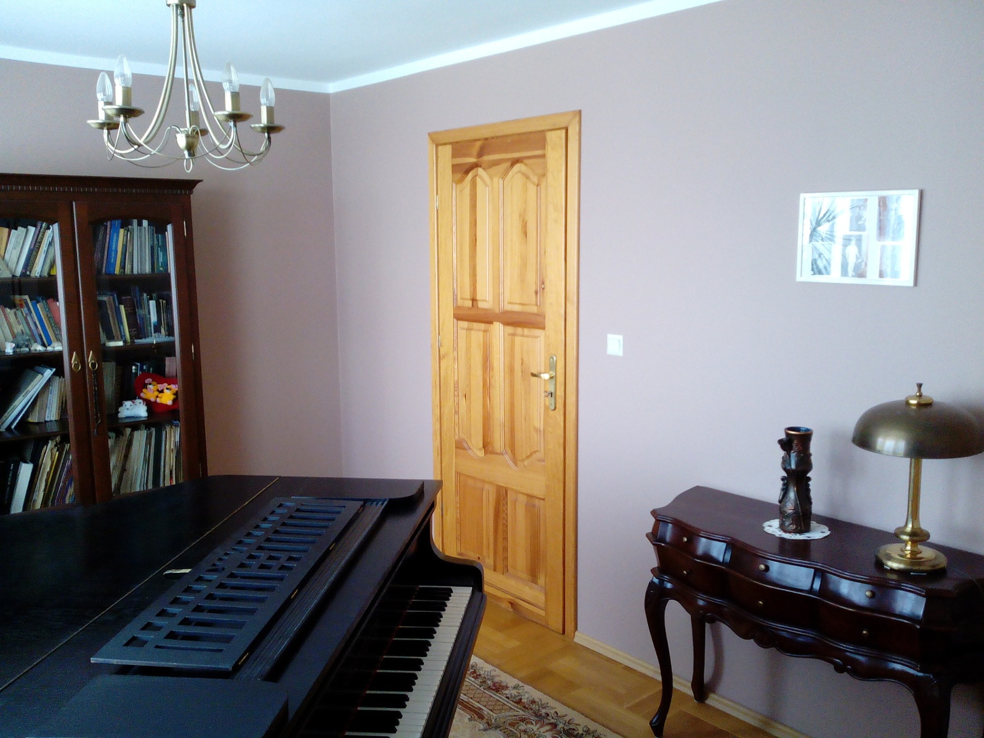 Jasny pokój z pianinem, drewnianą komodą i drzwiami z surowego drewna, ściany pomalowane na jasny odcień fioletu.