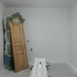 Przygotowanie do malowania pokoju: zabezpieczone meble pod folią, drewniane drzwi oparte o ścianę, oraz aluminiowy podest malarski na betonowej podłodze, świeżo pomalowane ściany na biało.