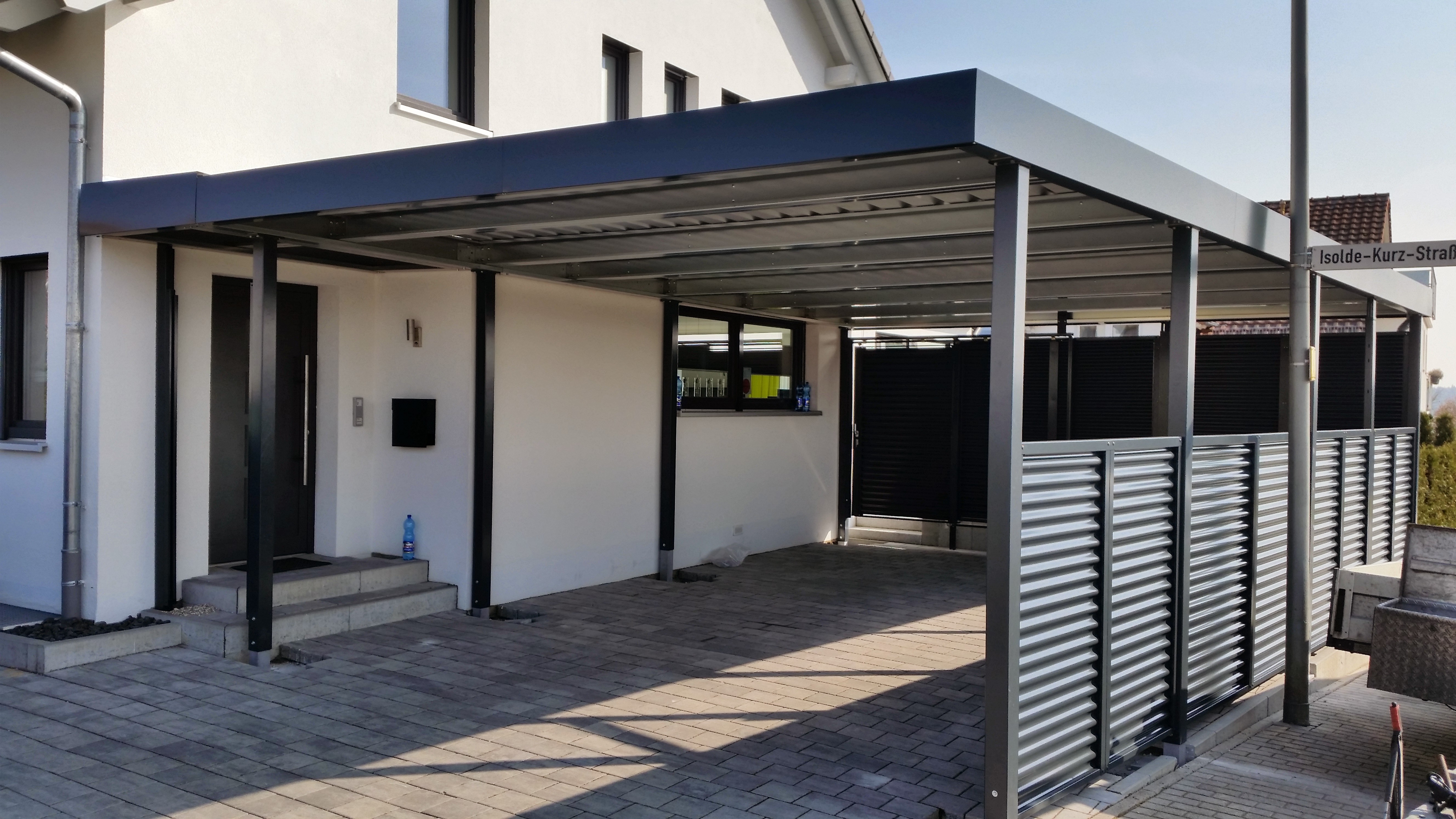 Nowoczesna wiata garażowa z metalową konstrukcją, przylegająca do białego budynku mieszkalnego, z widocznym fragmentem ogrodzenia z metalowych paneli żaluzjowych i kostką brukową na podjeździe.