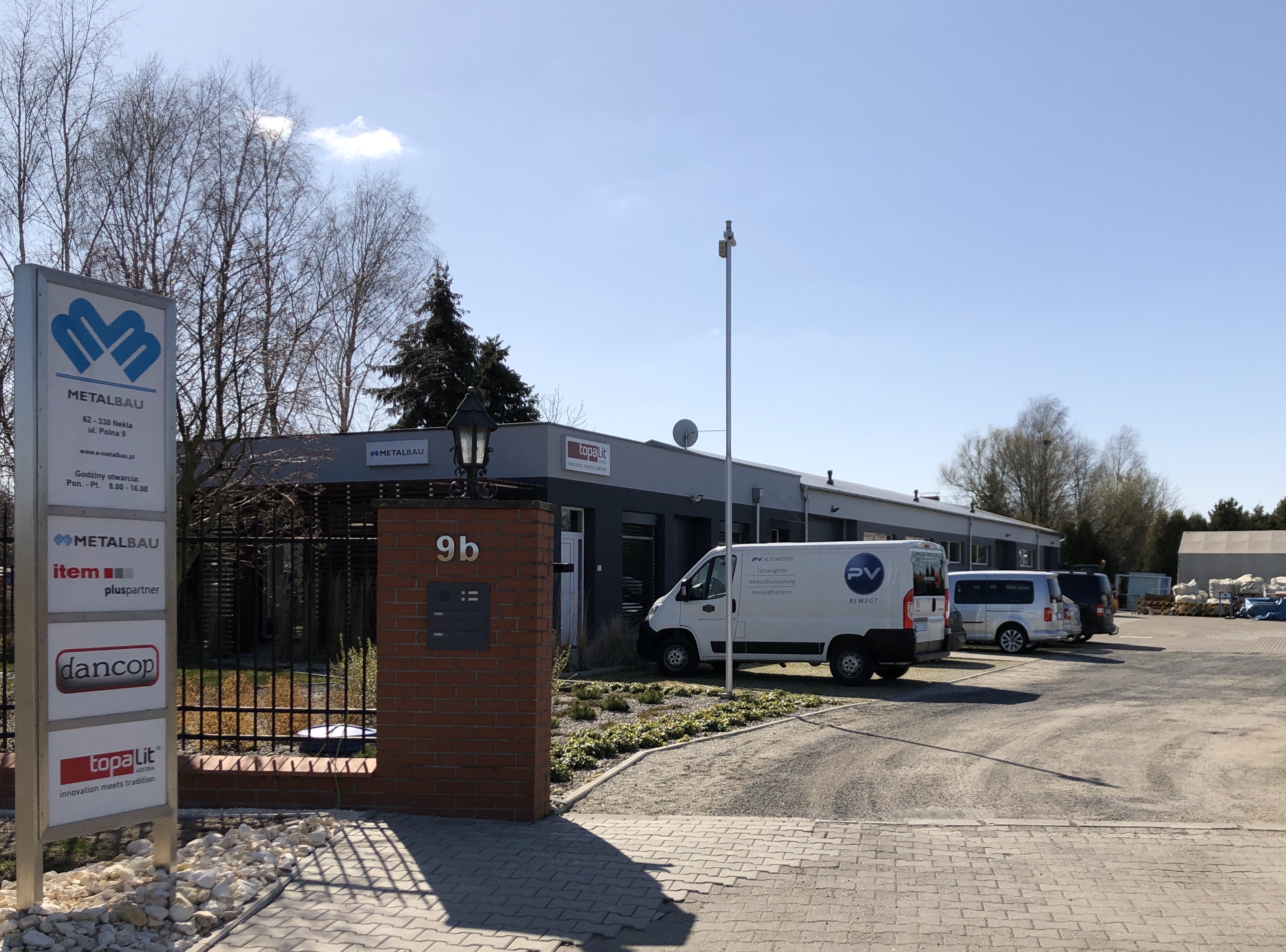 Budynek firmy Metalbau z widocznym logo, adresem, godzinami otwarcia, oraz zaparkowanymi samochodami dostawczymi PV Automotive na brukowanym podjeździe.