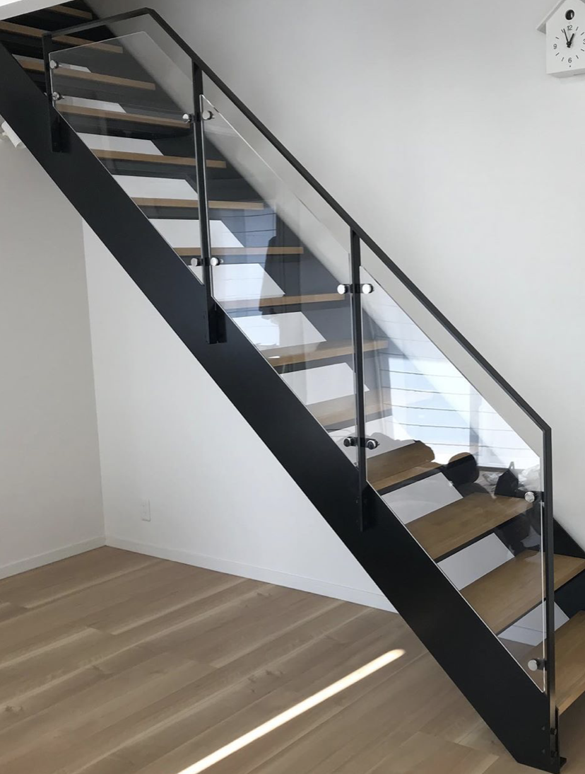 Nowoczesne schody metalowe z drewnianymi stopniami i szklaną balustradą mocowaną na okrągłych uchwytach, widok z dołu w minimalistycznym wnętrzu.