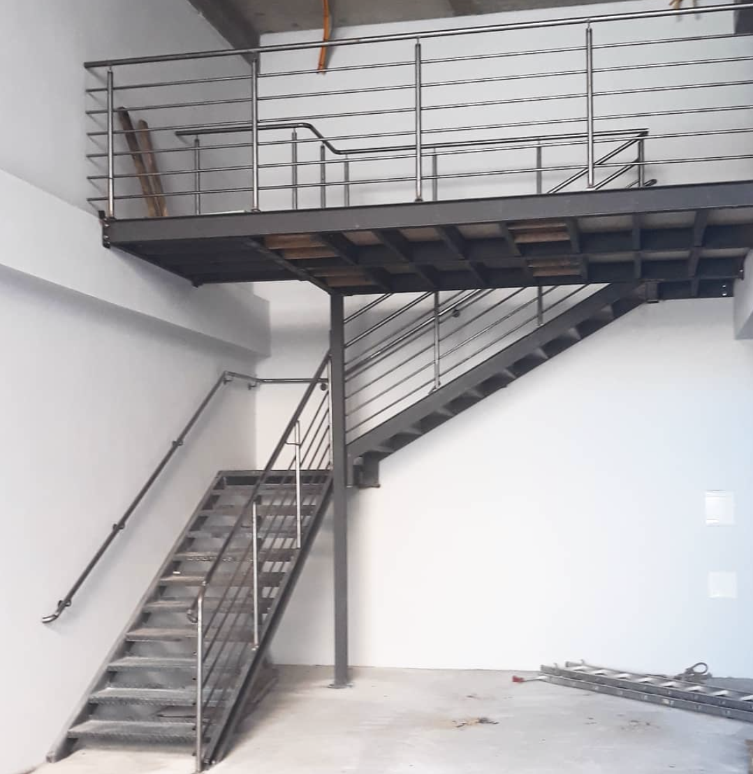 Metalowe schody z balustradą na antresolę w surowym wnętrzu, widoczne elementy konstrukcyjne i drabina w tle.