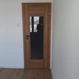 Dębowa drzwi wewnętrzne z czarnym szkłem wstawione w białą ścianę, widoczna klamka i zamek, na podłodze panele imitujące drewno.