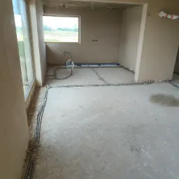 Surowe wnętrze w trakcie prac wykończeniowych: ściany po nałożeniu tynku, widoczne instalacje elektryczne i hydrauliczne wzdłuż ścian i pod oknem, surowy betonowy podkład podłogowy.