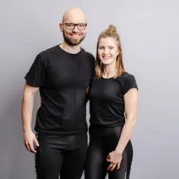 Uśmiechnięta para w czarnych strojach sportowych na szarym tle, mężczyzna w okularach obejmuje kobietę ramieniem.