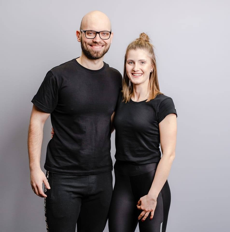 Uśmiechnięta para w czarnych strojach sportowych na szarym tle, mężczyzna w okularach obejmuje kobietę ramieniem.